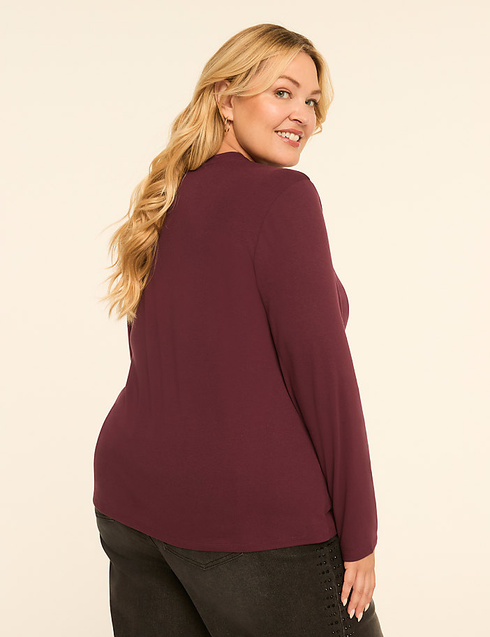 A woman with blonde hair wearing a maroon long-sleeved shirt and black pants smiles over her shoulder against a plain, light-colored background.