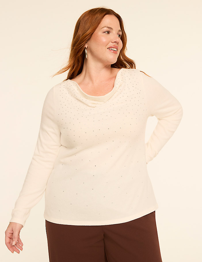 A woman with auburn hair smiles as she poses in a cream-colored, embellished cowl-neck sweater and brown pants against a plain, light background.