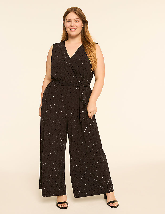 A woman in a black polka dot jumpsuit stands against a plain, light-colored background.