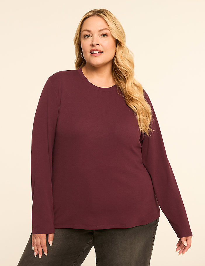 A blonde woman in a maroon long-sleeved shirt and dark jeans stands against a plain, light-colored background.