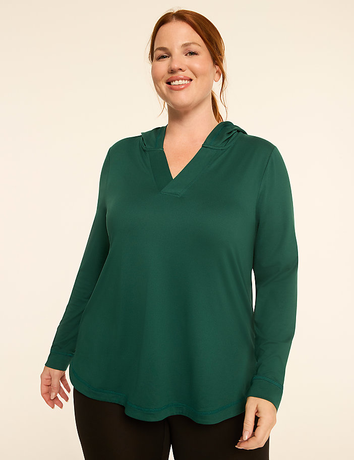 A smiling woman in a green hooded shirt stands against a plain, light-colored background.