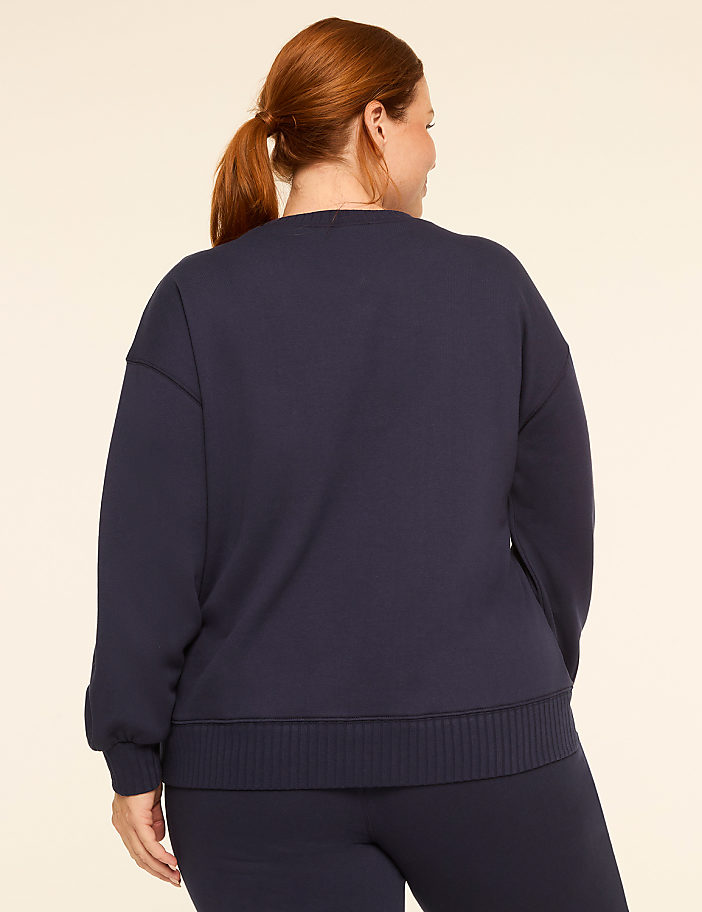A woman with auburn hair pulled into a ponytail stands facing away from the camera, wearing a navy blue sweatshirt and pants against a plain, light-colored background.