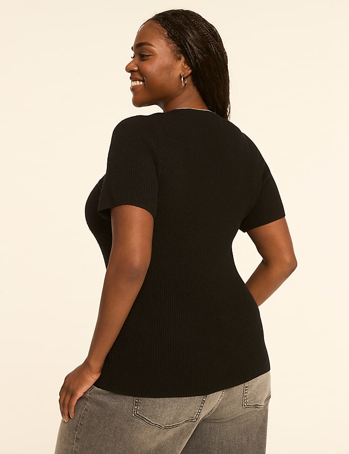A woman in a black ribbed top and grey jeans smiles over her shoulder against a plain, light-colored background.
