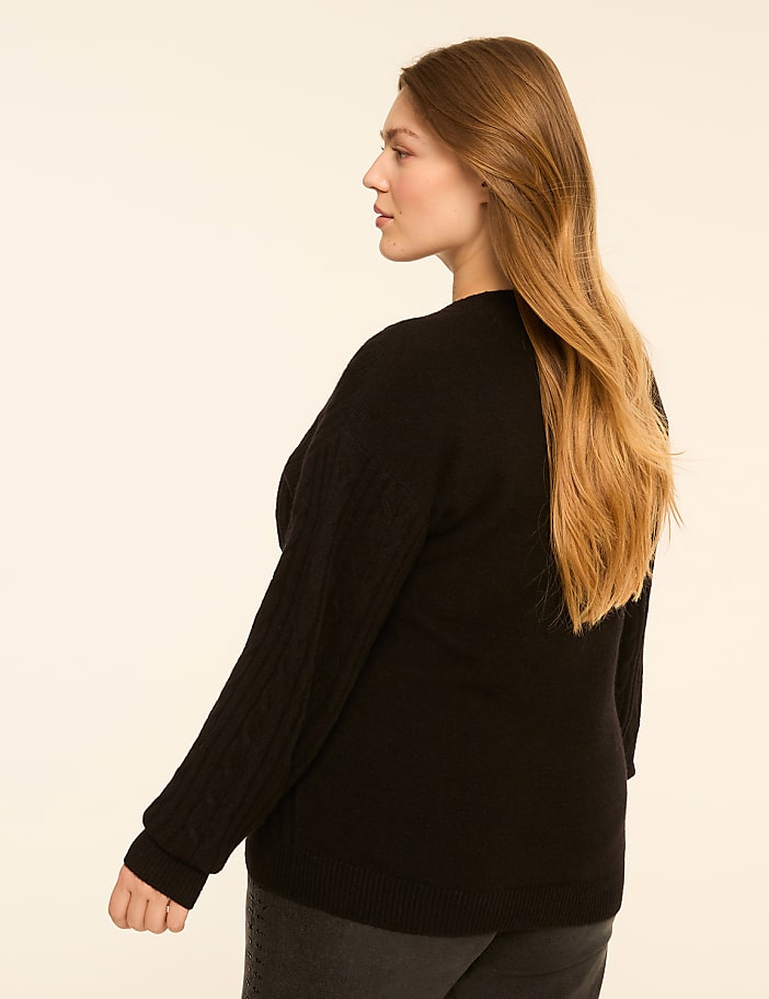 A woman in a black cable-knit sweater and dark jeans stands against a plain, light-colored background.
