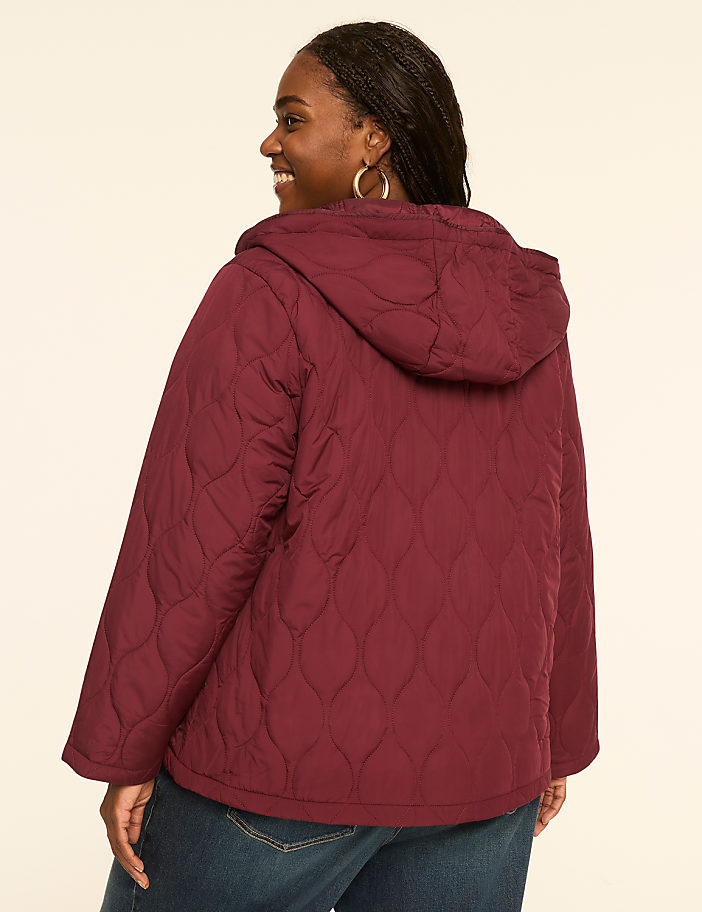 A woman in a maroon quilted jacket and blue jeans smiles over her shoulder against a plain, light-colored background.