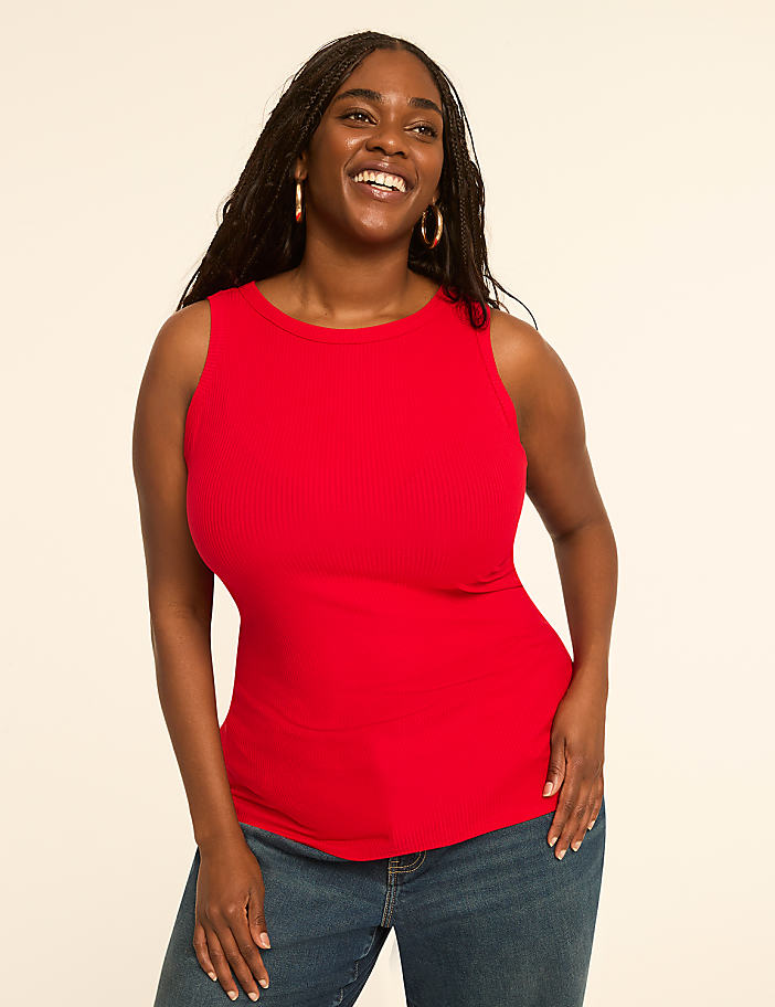 A smiling woman in a red tank top and blue jeans stands against a plain, light-colored background.