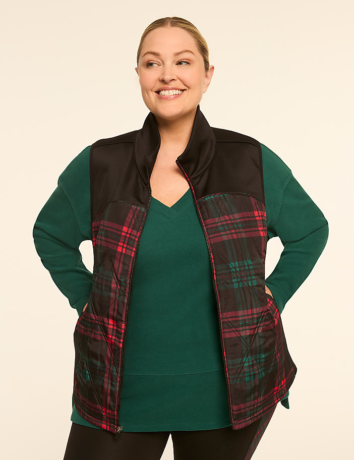 A smiling woman in a green sweater and a plaid vest stands against a plain, light-colored background.