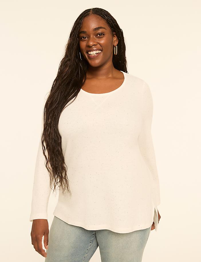 A smiling woman with long dark hair wears a white waffle-knit top and light blue jeans against a plain, light-colored background.