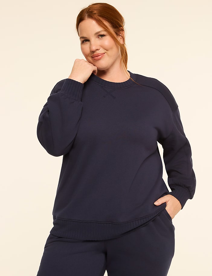 A woman in a navy sweatshirt and sweatpants poses with her hand on her chin against a plain, light-colored background.