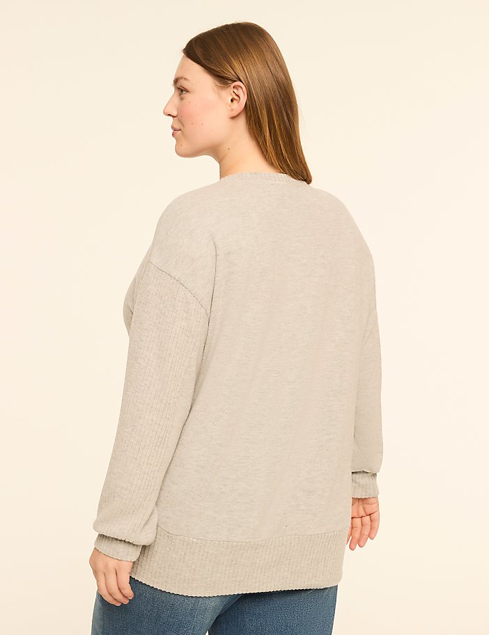 A woman in a light gray sweater and blue jeans stands against a plain, light beige background.
