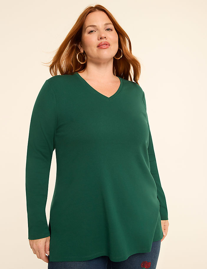 A woman with auburn hair and large hoop earrings poses in a green v-neck long-sleeved shirt against a plain, light-colored background.