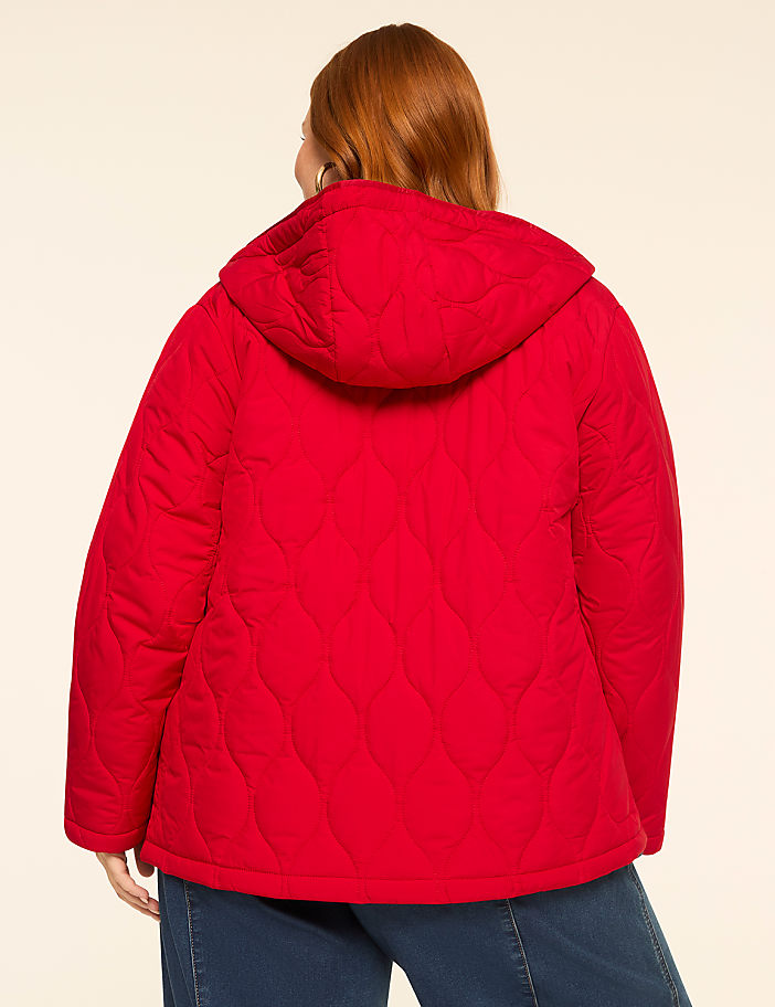 A person wearing a red quilted hooded jacket stands against a plain, light-colored background.