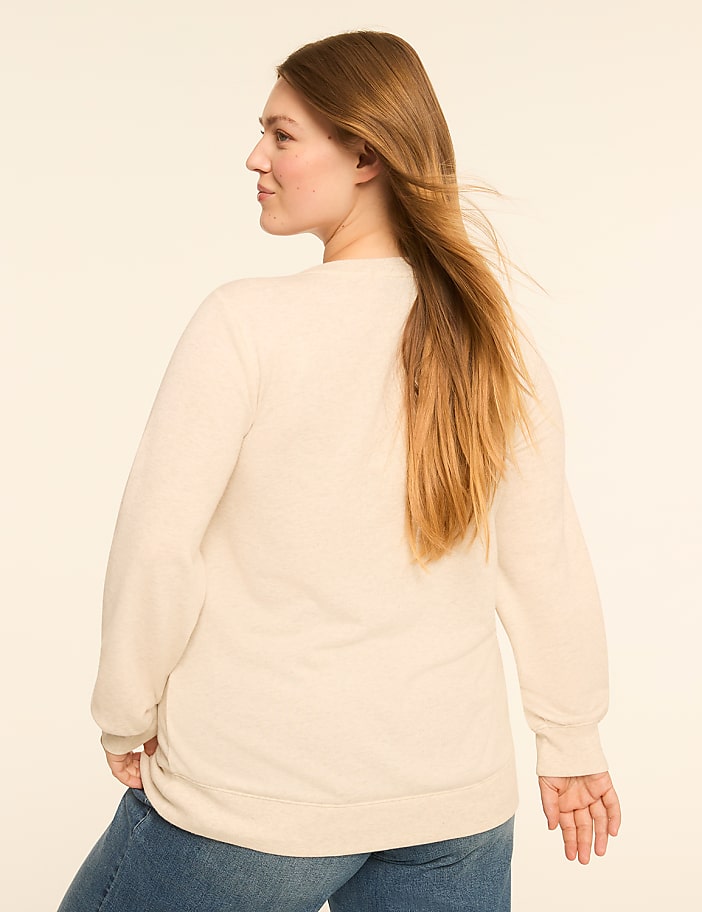 A woman with long blonde hair, wearing a cream-colored sweater and blue jeans, stands against a plain, light-colored background.