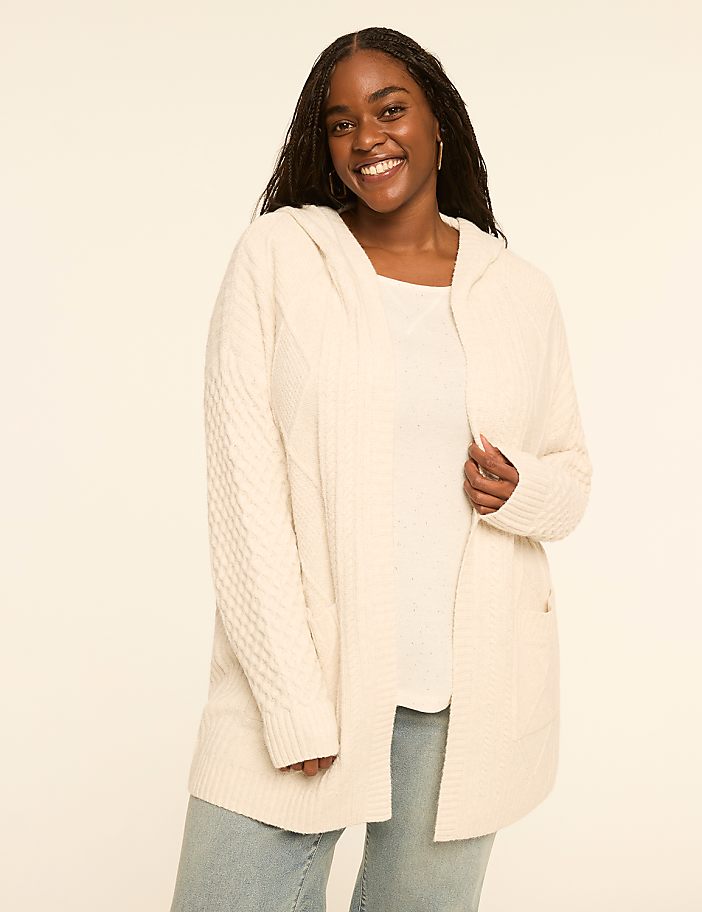 A smiling woman in a cream-colored hooded cardigan and light blue jeans stands against a plain, light background.