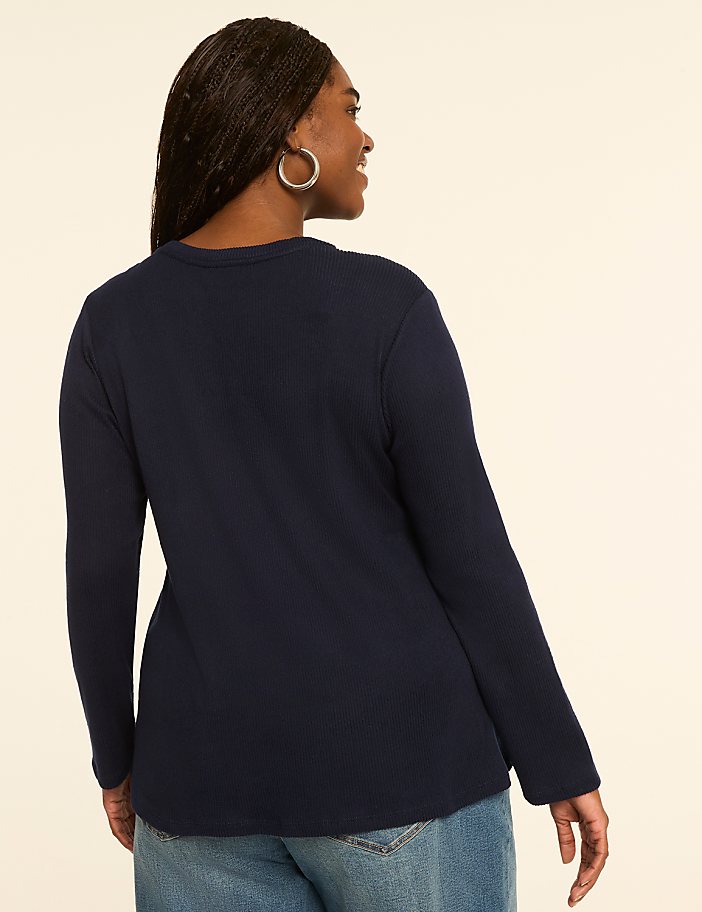 A woman in a navy long-sleeved shirt and jeans stands against a plain, light-colored background.
