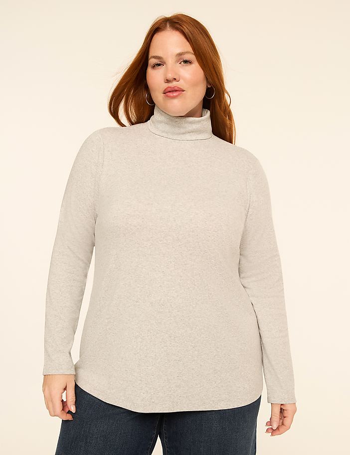 A woman with red hair and fair skin, wearing a light gray turtleneck sweater and dark blue jeans, stands against a plain, light beige background.
