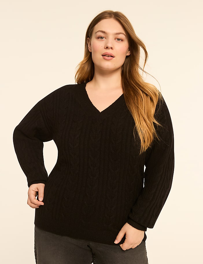A woman in a black cable-knit sweater stands against a plain, light-colored background.