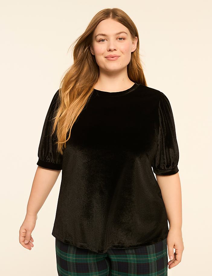 A woman with long blonde hair smiles at the camera, wearing a black velvet top and plaid pants against a plain background.