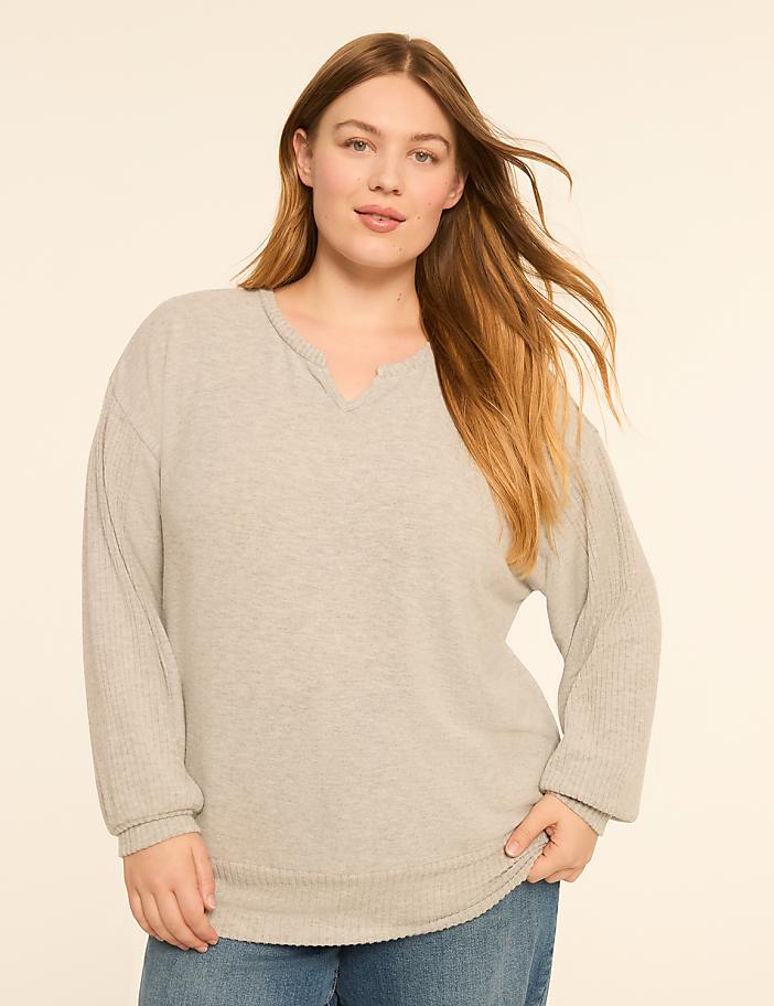 A woman with long, blonde hair stands in the foreground wearing a light gray, long-sleeved sweater and blue jeans, against a plain, light beige background.