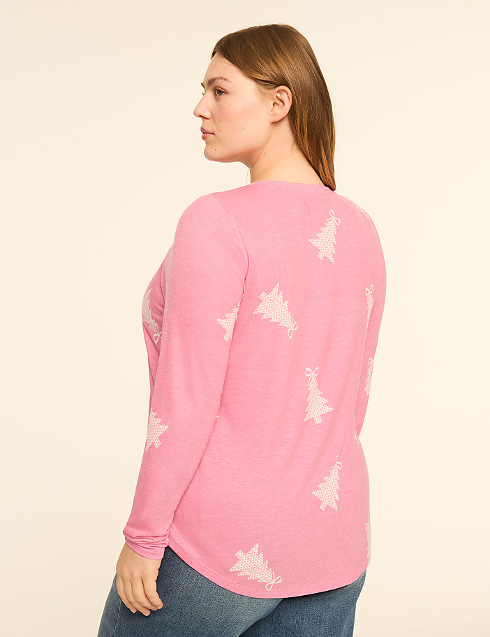 A woman in a pink long-sleeved shirt with a Christmas tree pattern stands against a plain, light-colored background.