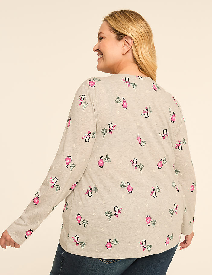 A woman in a light gray long-sleeved shirt with a repeating pattern of pink penguins and Christmas trees smiles over her shoulder against a plain, light background.