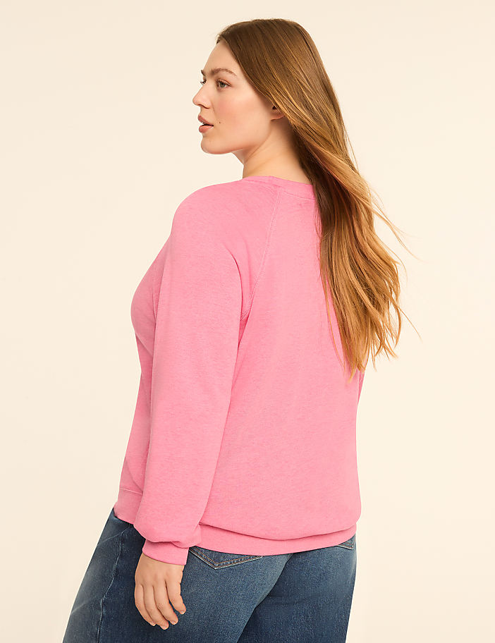 A woman in a pink sweatshirt and blue jeans stands against a plain, light-colored background.