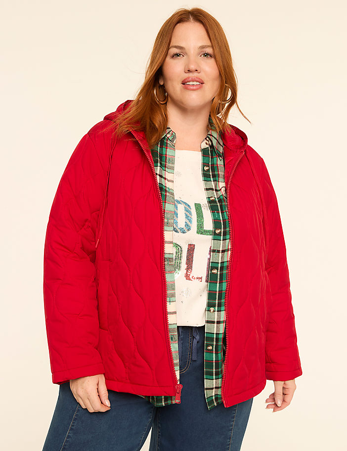 A woman in a red quilted jacket and plaid shirt stands against a plain, light-colored background.