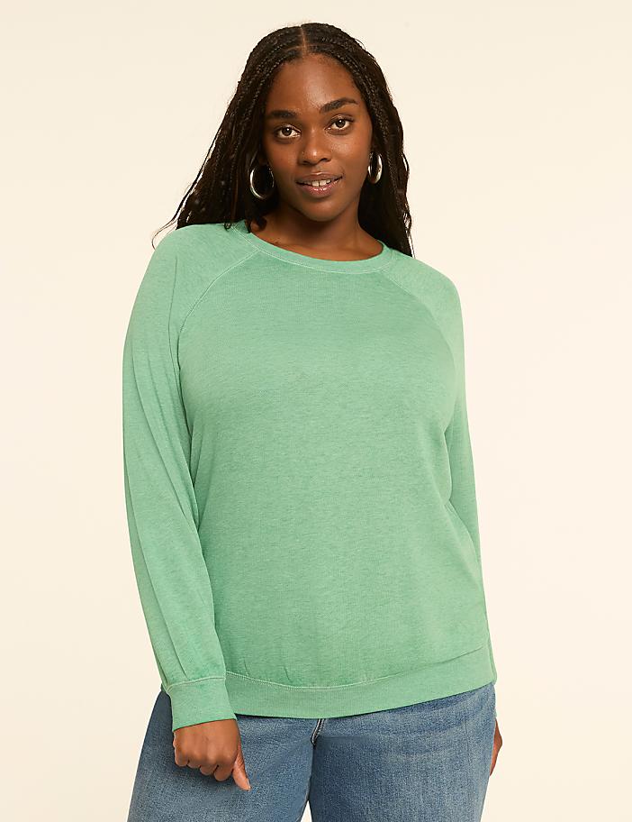 A woman with dark braided hair and hoop earrings, wearing a light green long-sleeved shirt and blue jeans, stands against a plain, light-colored background.