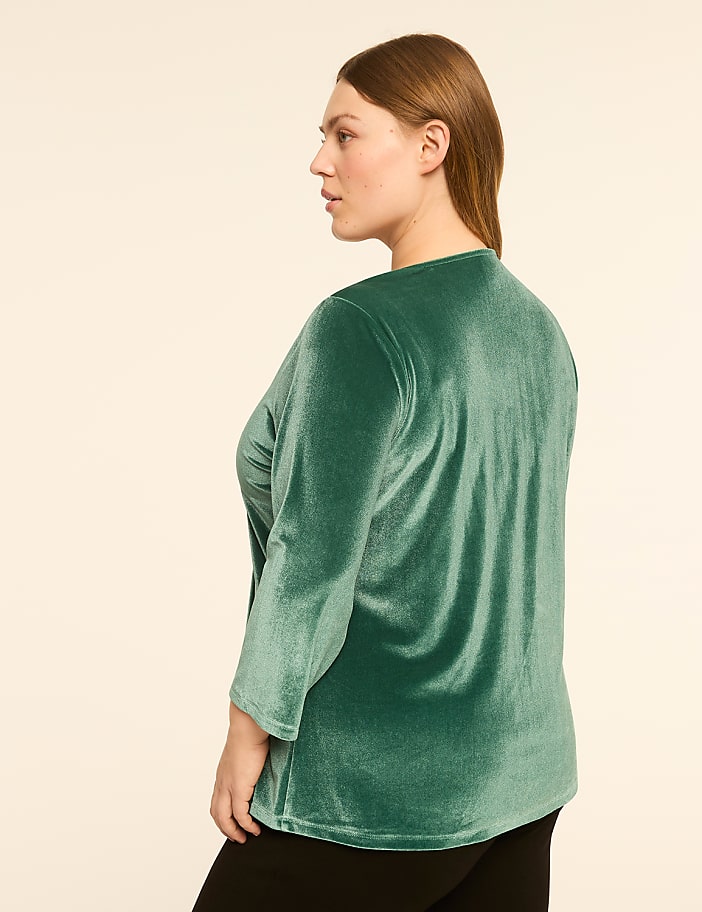 A woman in a green velvet top stands against a plain, light-colored background.