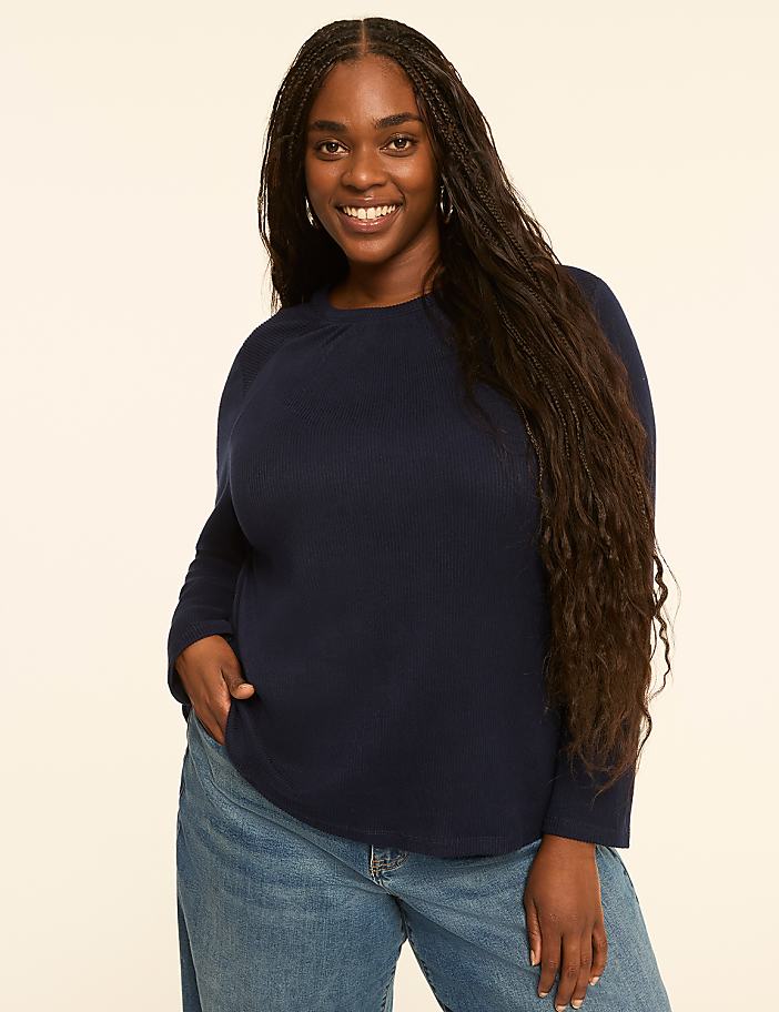 A smiling woman with long dark hair and a navy blue waffle-knit shirt stands against a plain, light-colored background.