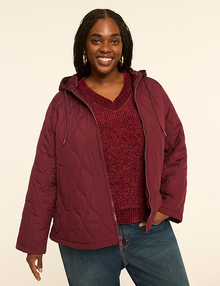 A smiling woman in a quilted burgundy jacket and a v-neck sweater stands against a plain, light-colored background.