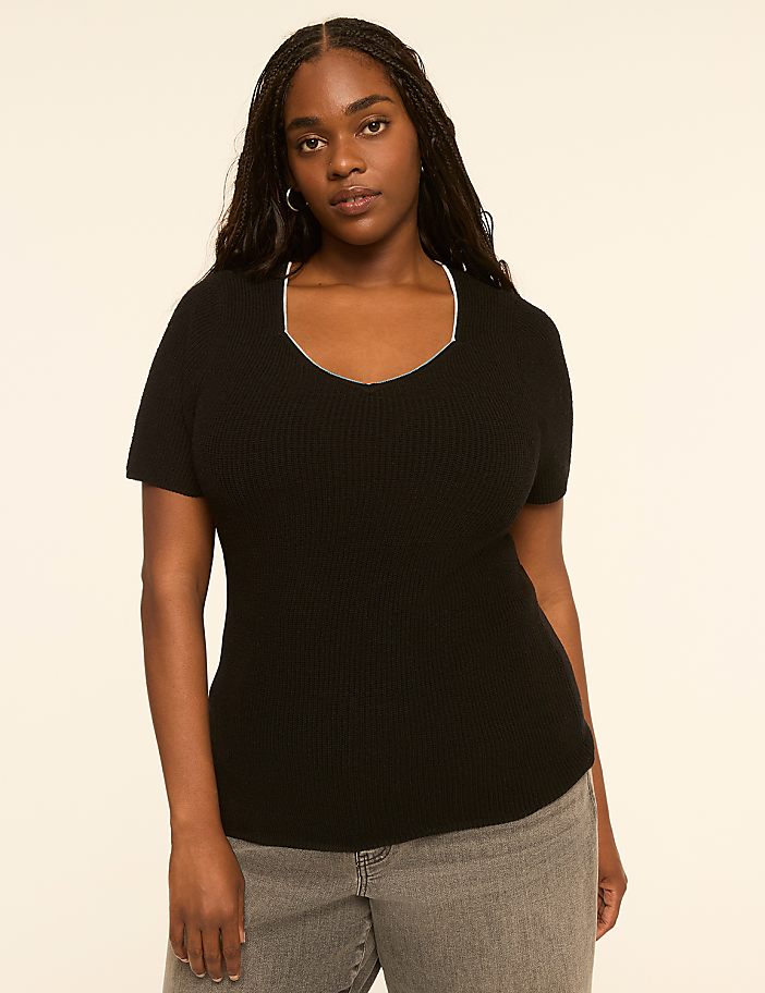 A woman in a black v-neck shirt and grey jeans stands against a plain, light-colored background.