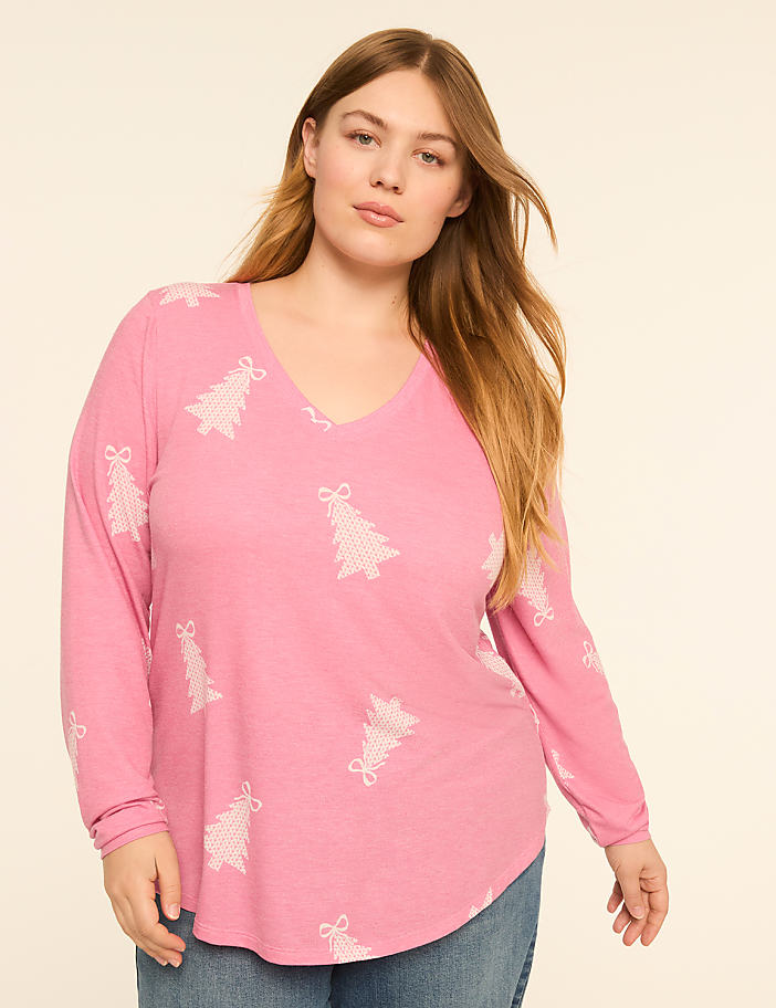 A woman in a pink long-sleeved shirt with a Christmas tree pattern stands against a plain, light-colored background.