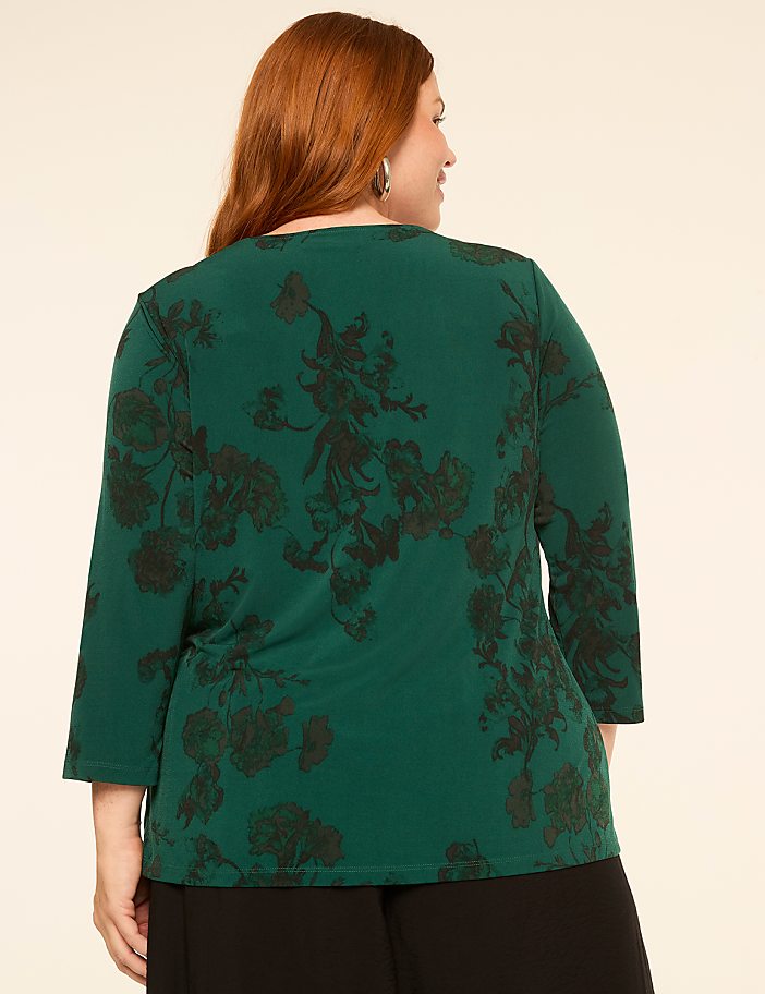 A woman with red hair wearing a green floral top stands against a plain beige background.