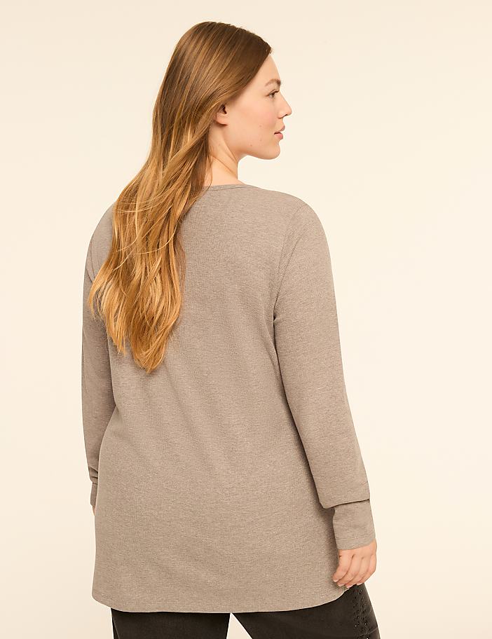 A woman with long, blonde hair stands facing away from the camera in a beige waffle-knit top and dark pants against a plain, light-colored background.