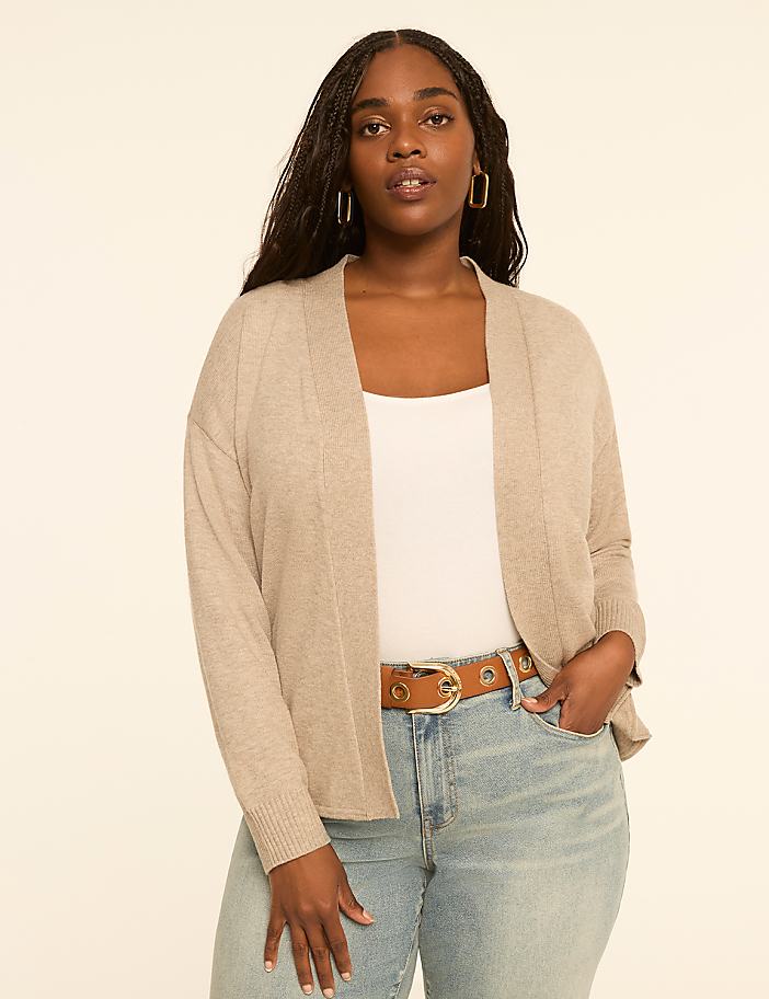 A woman in a beige cardigan and jeans stands against a plain, light-colored background.