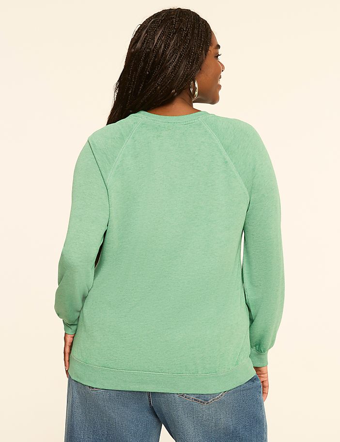 A woman in a green sweatshirt and blue jeans stands against a plain, light-colored background.
