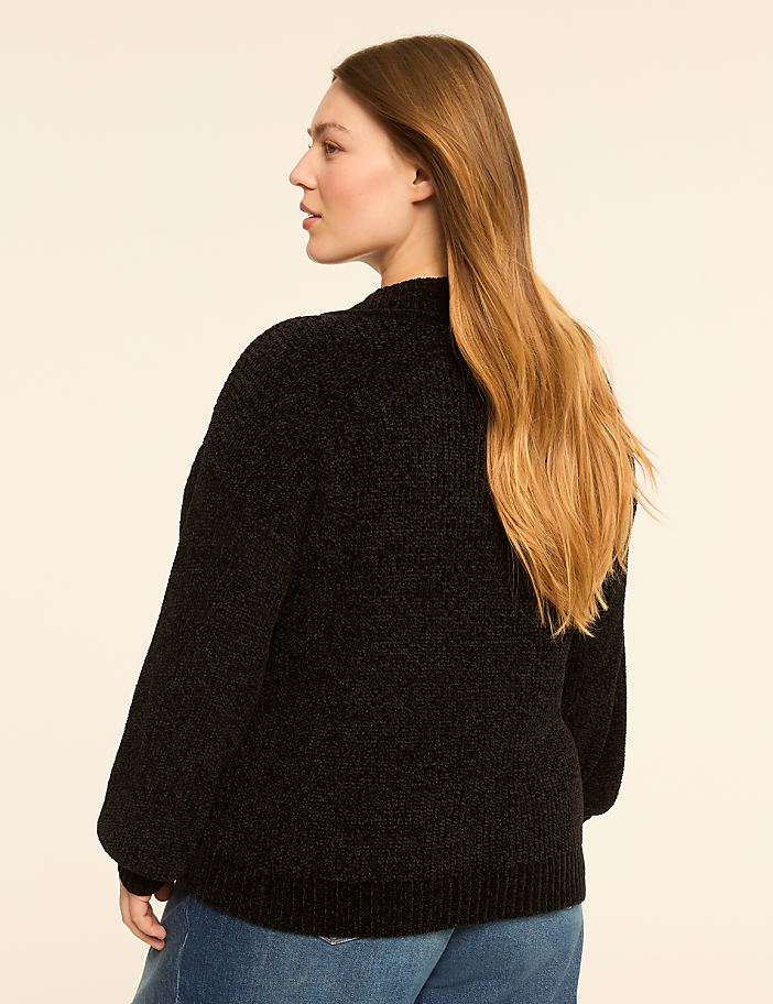 A woman in a black sweater and blue jeans stands against a plain, light-colored background.