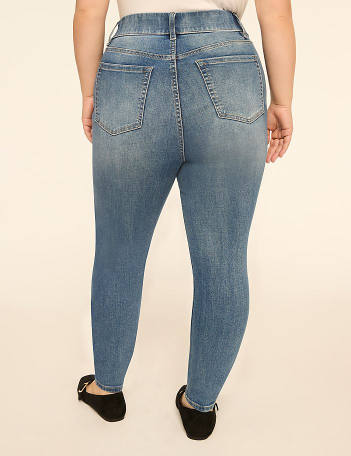 A person wearing blue denim jeans and black flats stands against a plain, light-colored background.