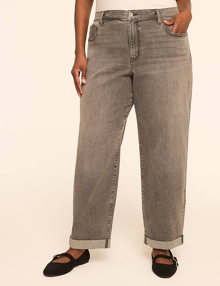 A person stands in the foreground wearing cuffed grey jeans and black ballet flats against a plain, light-colored background.