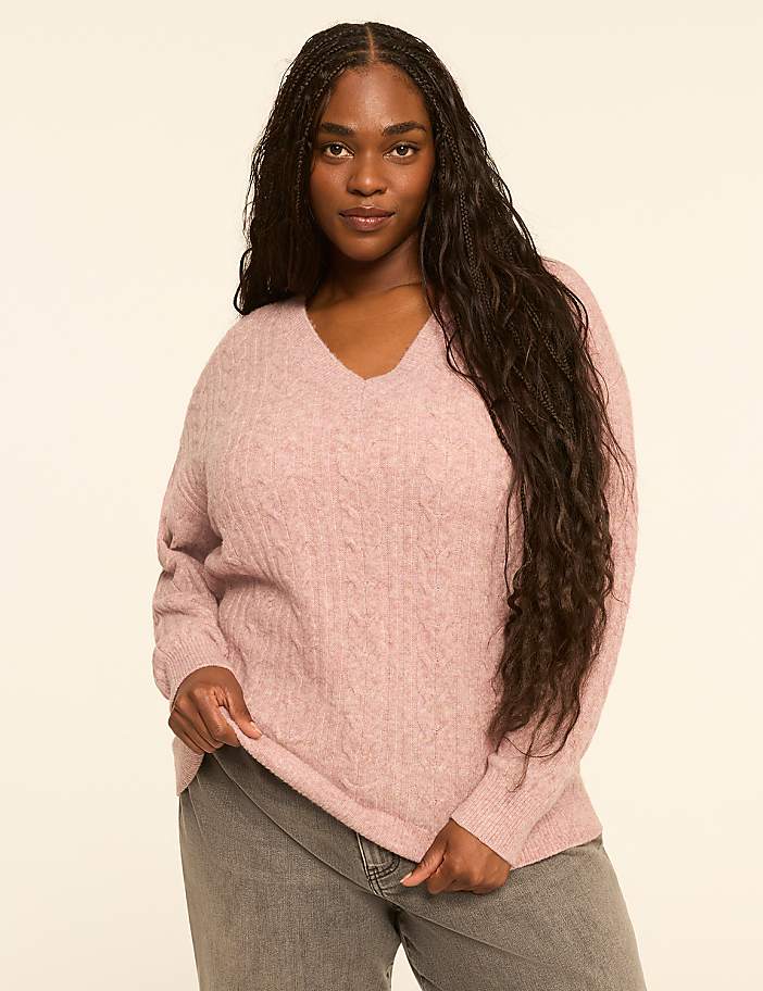 A woman with long, dark hair stands in the foreground wearing a pink cable-knit sweater and gray jeans against a plain, light-colored background.