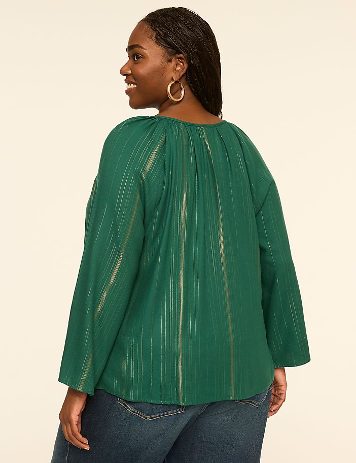 A woman in a green, metallic-striped blouse and blue jeans stands against a plain, light-colored background.