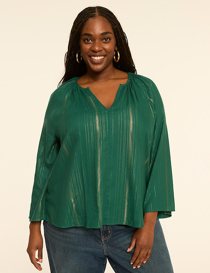 A smiling woman with dark hair and hoop earrings, wearing a green, metallic-striped blouse and blue jeans, stands against a plain, light-colored background.