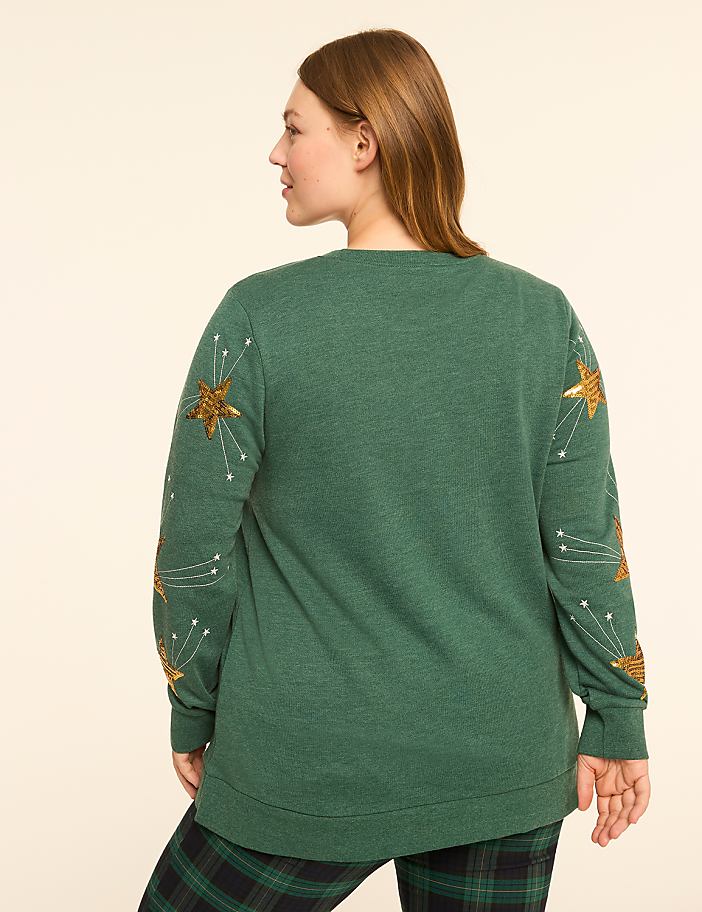 A woman in a green sweater with gold star embellishments on the sleeves stands against a plain, light-colored background.