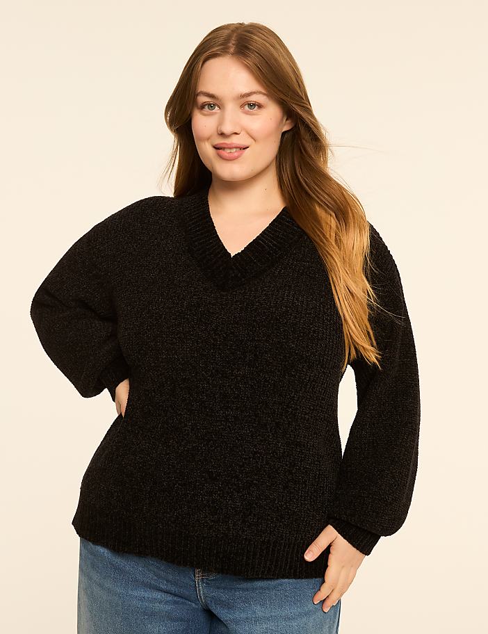 A woman in a black sweater and blue jeans stands against a plain, light-colored background.
