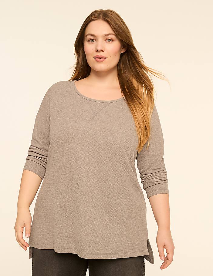 A woman with long brown hair poses in a taupe waffle-knit top against a plain, light-colored background.