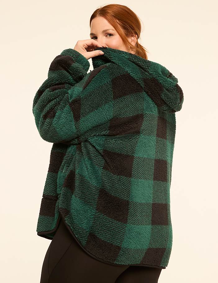 A woman in a green and black plaid fuzzy jacket peeks over her shoulder against a plain cream background.