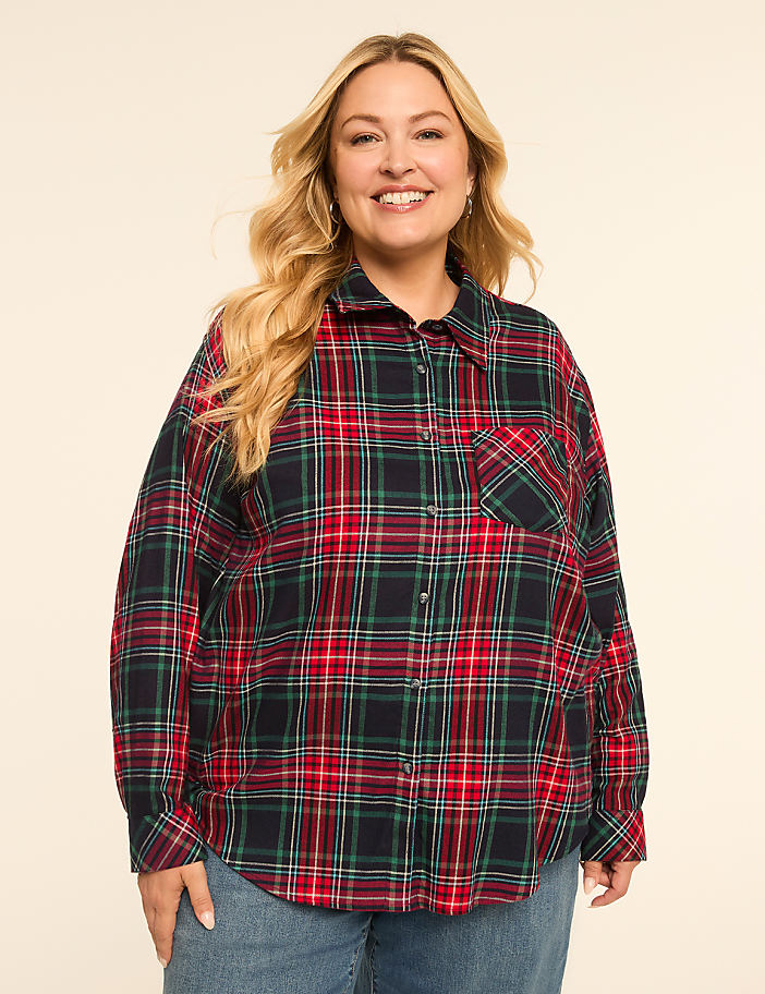 A smiling woman with blonde hair wears a red, green, and blue plaid shirt and blue jeans against a plain beige background.