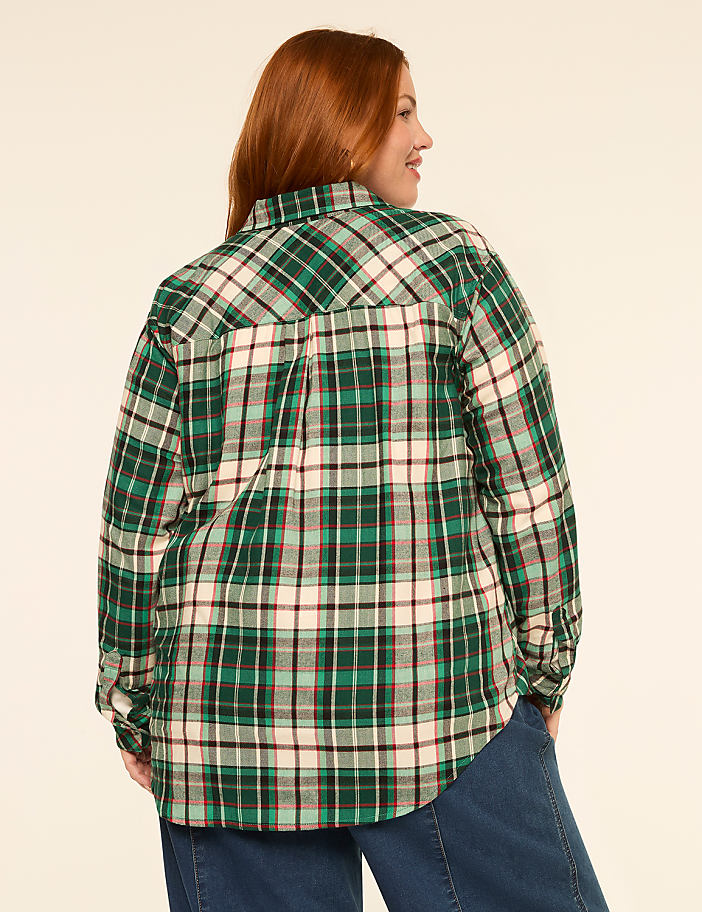 A woman with red hair wearing a green plaid shirt stands against a plain, light-colored background.