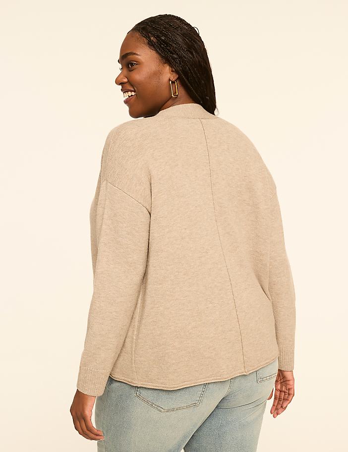 A woman in a beige sweater and jeans smiles over her shoulder against a plain, light-colored background.